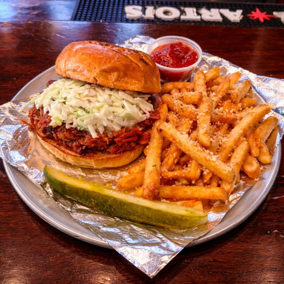 Pulled pork sandwich with coleslaw, a pickle spear, and parmesan fries with ketchup on a metal plate.